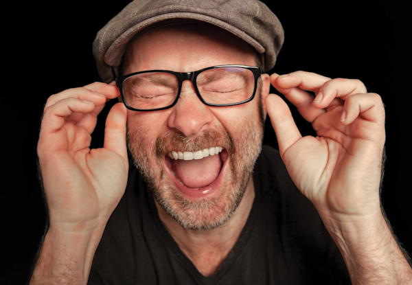 Man in cap laughing and holding glasses, black background.