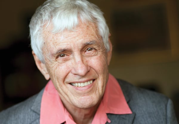 Elderly man smiling warmly, wearing a grey jacket and pink shirt.