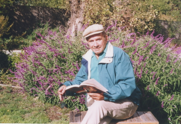 Elderly man reading a book outdoors, sitting by purple flowers.