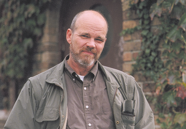 Bald man with beard in green jacket standing before ivy-covered wall.