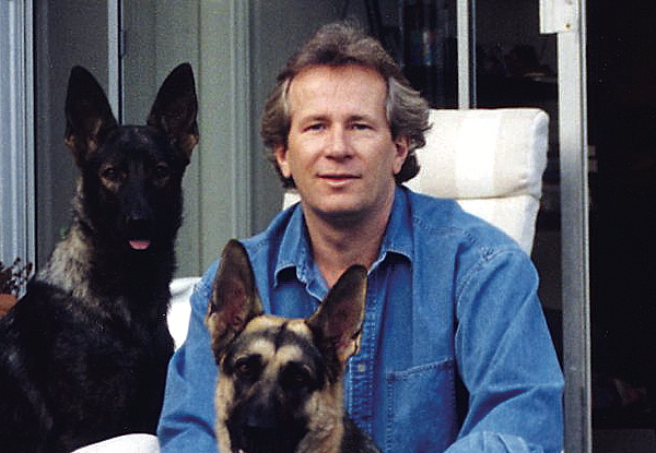 Man in a denim shirt sitting with two German Shepherds.