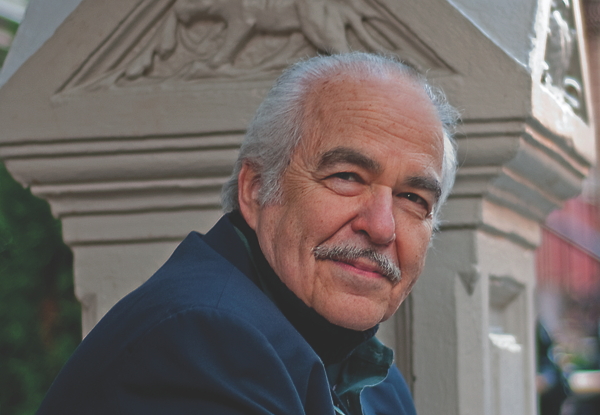 Elderly man with a mustache smiling near a column.