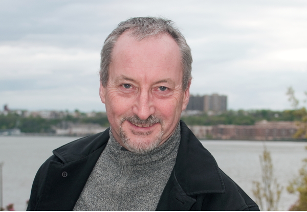 "Smiling older man with a beard by a riverfront on a cloudy day."