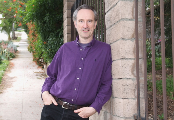 Man in purple shirt standing by a stone pillar outside.
