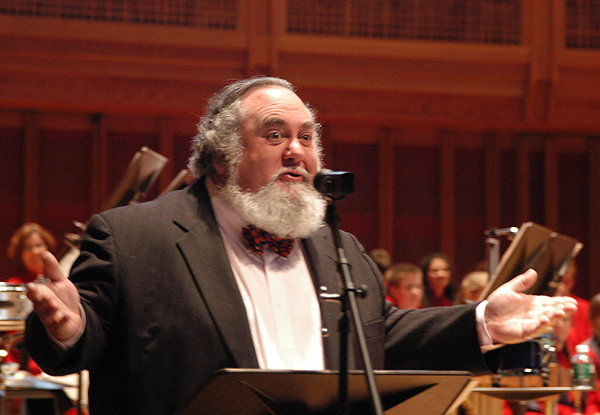 Conductor speaking into a microphone with orchestra in background.