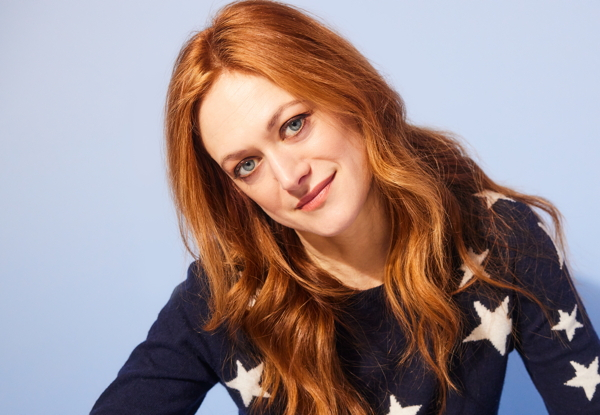 Woman with red hair smiling in a star-patterned sweater, blue background.