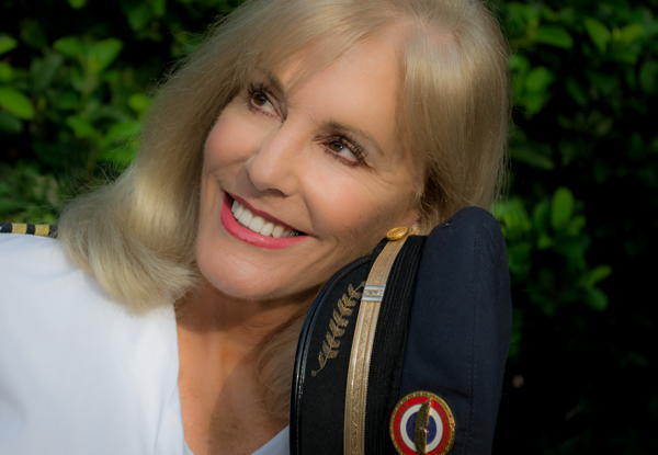 Elderly woman smiling, holding a pilot's cap, with leafy background.