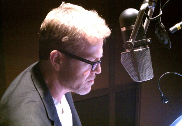 Man in glasses recording in a studio with a microphone.