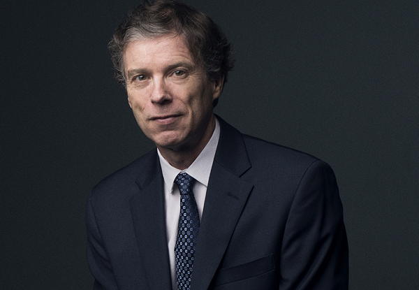 Mature man in suit with subtle smile against dark background.