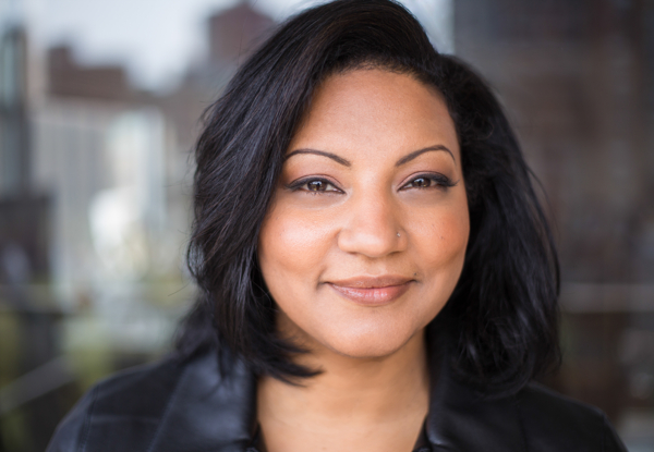 Portrait of a smiling woman with short black hair, wearing a leather jacket. Cityscape in background.
