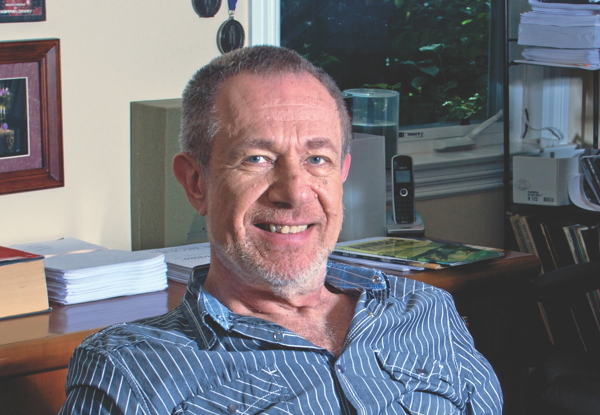 Man smiling in striped shirt, seated at a desk with papers and a phone.