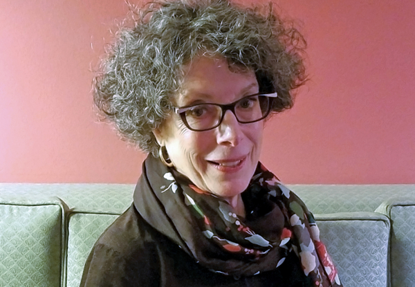 Woman with curly gray hair and glasses smiling, wearing a scarf, seated on a couch.