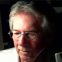 Elderly man with glasses looking down, low-light setting.