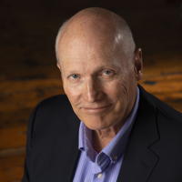 Portrait of a bald man in a suit and purple shirt, smiling against a wooden background.