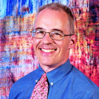 Smiling man in glasses with a blue shirt, in front of a colorful backdrop.
