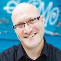 Smiling bald man with glasses against a blue graffiti wall.
