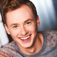 Smiling young man with short blonde hair in a grey patterned shirt.
