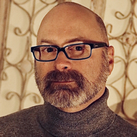 Bald man with beard, glasses, and turtleneck, with patterned background.