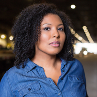 Portrait of a woman in a blue shirt with curly hair, blurred lights in the background.
