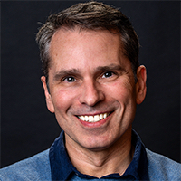 "Headshot of a smiling middle-aged man with short hair on a dark background."