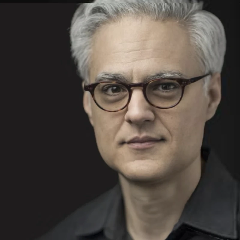 Portrait of a middle-aged man with gray hair and glasses on a black background.