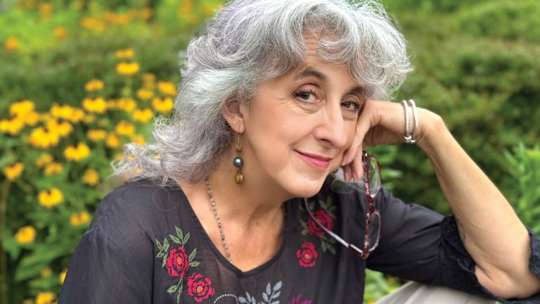 Elderly woman smiling in a garden, wearing floral embroidered top.